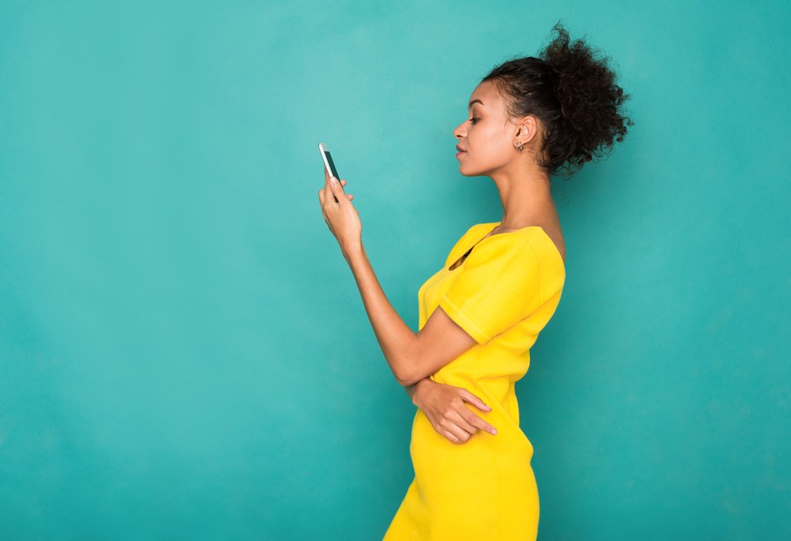 Young african-american woman typing message on smartphone on blue studio background, side view. Communication concept