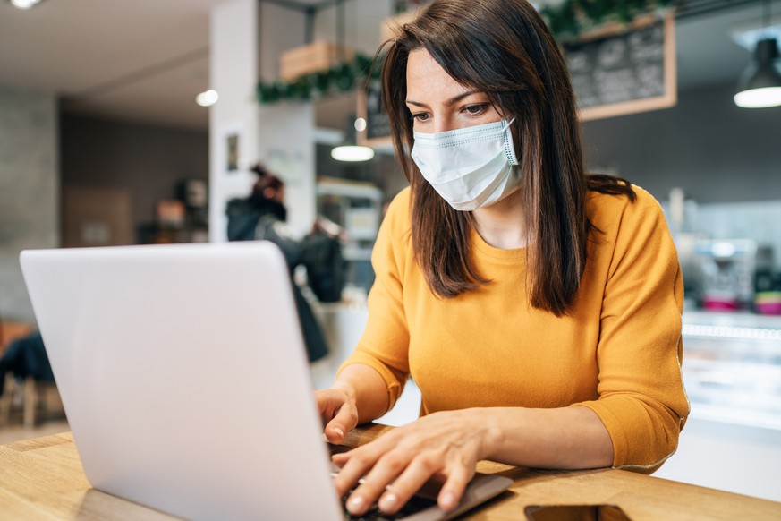 Portrait of young woman using laptop at cafe wearing face protective mask to prevent infectious diseases