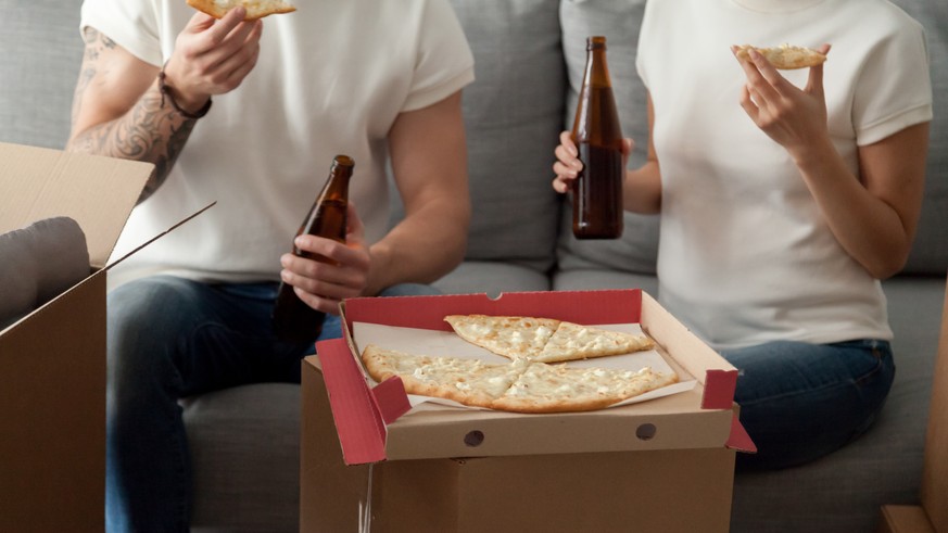 Couple eating pizza celebrating housewarming party on moving day, man and woman enjoying beer and snack sitting on sofa with unpacked boxes around, move in new home, delivery service, close up view, P ...
