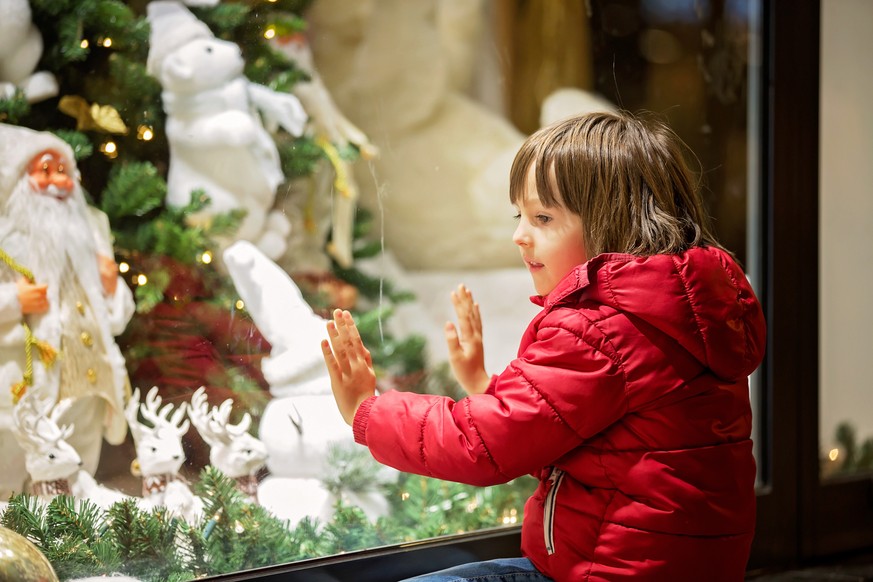 Beautiful little child, boy, watching Christmas decoration with toys in a shop window display, wishing for a present, his reflection in the window