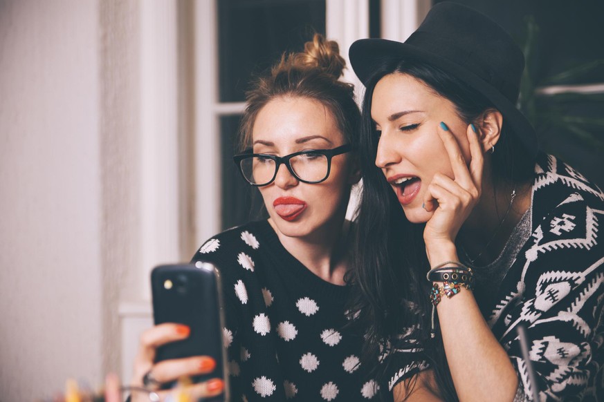 Vintage toned portrait of two fashionable young women taking a selfie photograph with a smartphone, sticking tongue out.