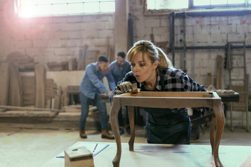 Portrait view of a female industrial worker in her 40s working as a carpenter in her workshop with sandpaper while her colleagues are in the background