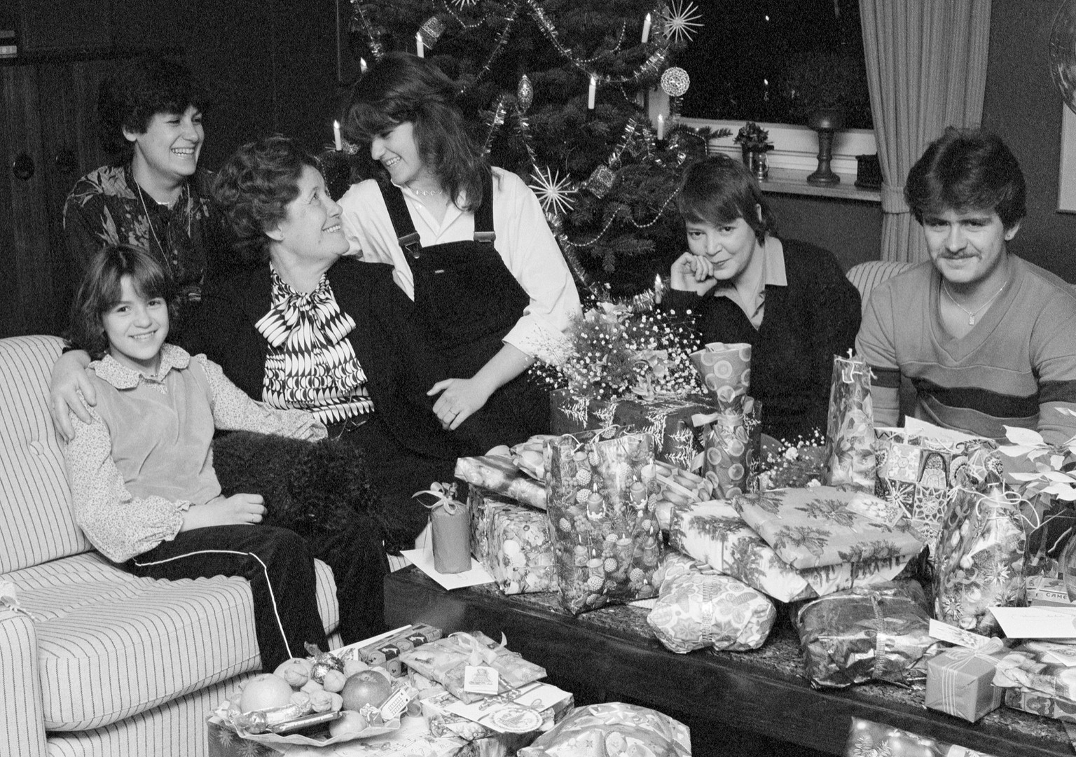 Siebziger Jahre, Familie sitzt am Heiligabend im Wohnzimmer zusammen und freut sich ueber viele Weihnachtsgeschenke auf dem Tisch Siebziger Jahre, Schwarzweissfoto, Feiertage, Weihnachten, Heiligabend ...