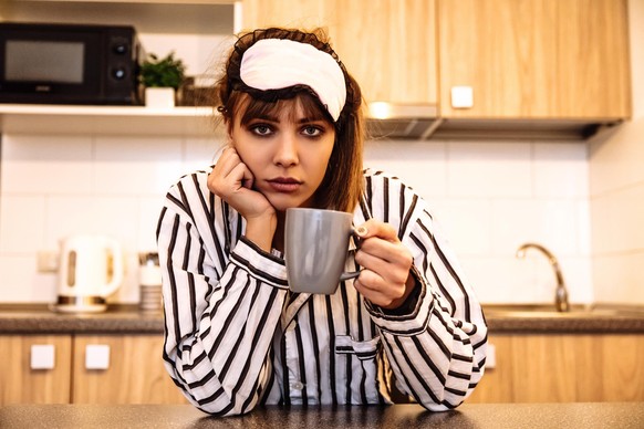 Early Morning. Girl in pajama and sleep mask is holding a cup and looking at camera, in the kitchen at home