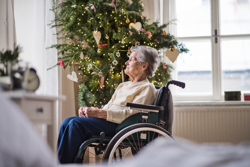A lonely senior woman in wheelchair at home at Christmas time, looking out of a window.