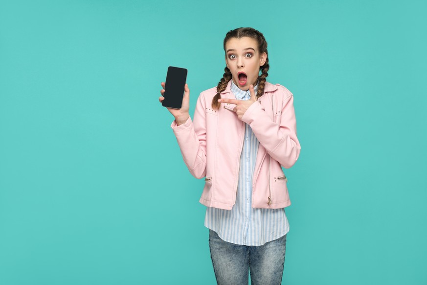 shocked funny girl in casual or hipster style, pigtail hairstyle, standing, holding and pointing at mobile display, screen with surprised face, Indoor studio shot, isolated on blue or green background