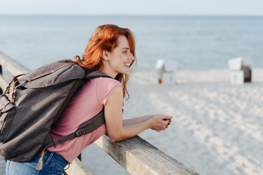 Young woman hiker or backpacker at the beach standing leaning over a wooden railing on a pier overlooking the sand with a smile