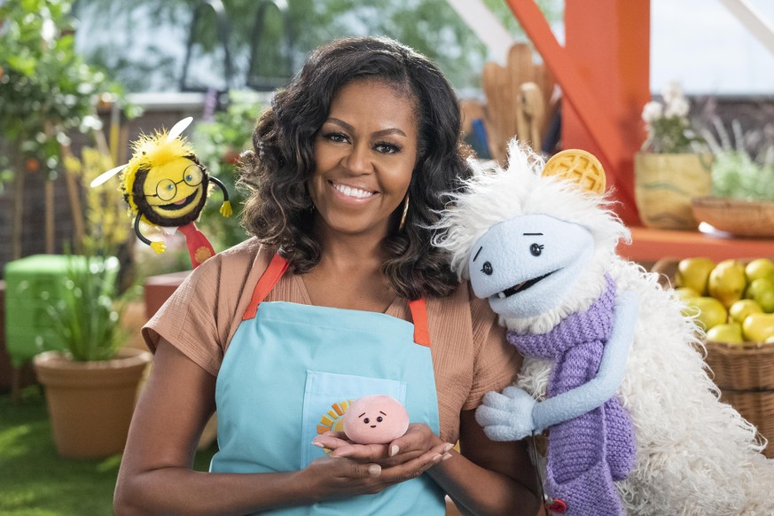Michelle Obama, wearing a blue apron, stands in a rooftop garden posing for the camera and holding a pink, round mochi puppet. A bee puppet wearing a red tie hovers over her shoulder while a furry whi ...