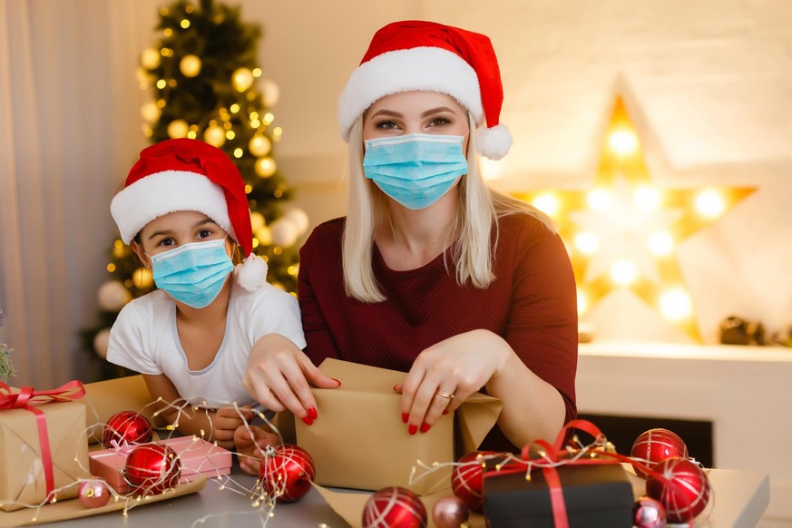Beautiful mother and daughter in medical masks have fun at home near the Christmas tree in a white interior. Family happiness, holiday, joy, vacation, games with a woman. New Year&#039;s preparations. ...