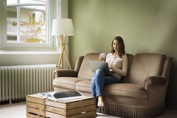 Young woman sitting on couch with cup of coffee using laptop model released Symbolfoto property released PUBLICATIONxINxGERxSUIxAUTxHUNxONLY MOEF00186