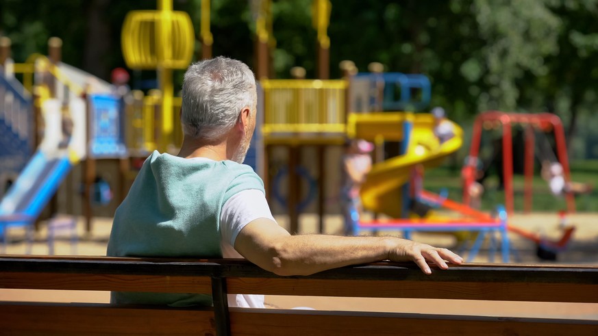 Male pensioner sitting on bench and watching grandkids on playground, family
